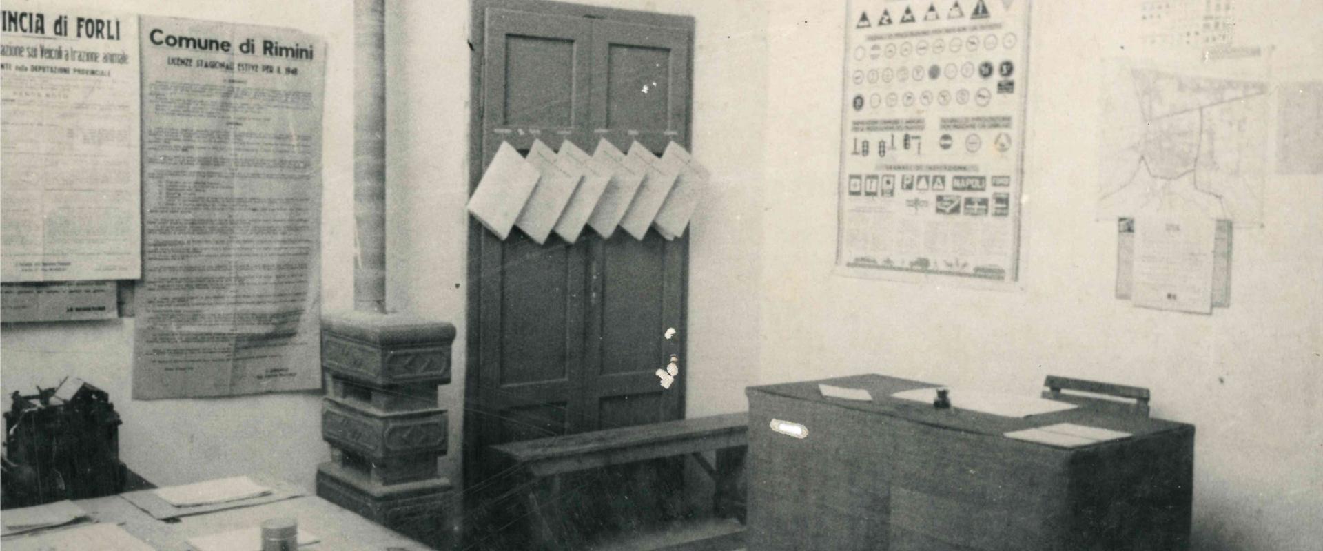 foto di un ufficio dei Vigili Urbani all'interno del palazzo Ceschina. Vi sono due scrivanie opposte con sopra timbri e carte, alle pareti dei manifesti tra cui uno del comune per le licenze estive 1948, l'altro con la pianta della città 