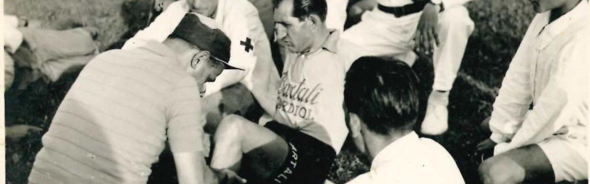 Immagine con al centro il ciclista Gino Bartoli mentre, a seguito di una caduta, veniva soccorso da alcuni medici. Attorno a lui alcune persone tra le quali il Vigile Urbano Sergio Ranchi in divisa bianca   