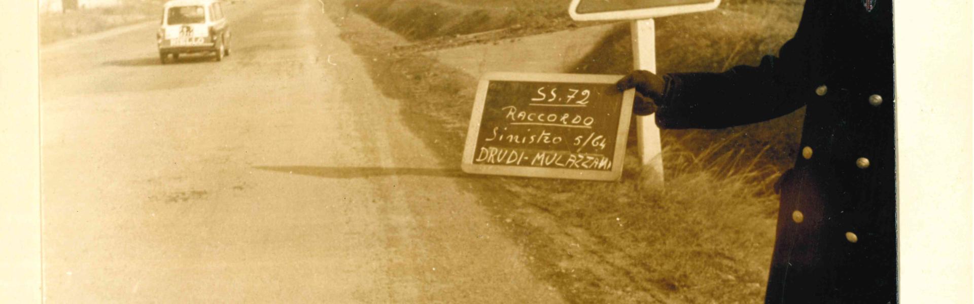 Riproduzione di una pagina dei un rapporto fotografico del 1964, con in primo piano un Vigile con in mano una lavagna indicante SS 72 raccordo, sullo sfondo un auto e i palazzi che ora sono in via Dalla Chiesa