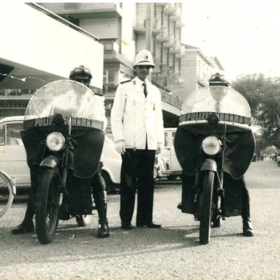 Immagine del Vigile Mondaini in divisa bianca e nera in mezzo a due motociclisti in piazzale Benedetto. Sullo sfondo via Pascoli, le terrazze dell'hotel Admiral