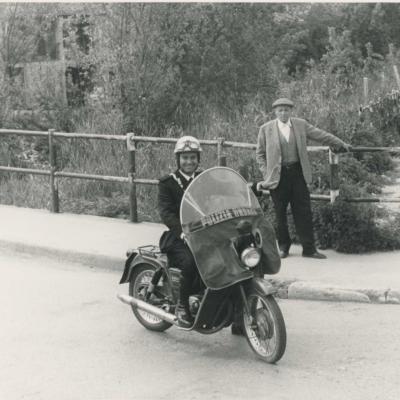 Il Vigile Urbano Painelli a sella alla moto Guzzi di servizio su un ponte. Sullo sfondo il torrente Ausa con la vegetazione non curata. Un anziano è appoggiato alla balaustra del ponte