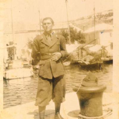 1942 Umberto Tamburini in piedi in divisa sulla banchina del porto dell'isola di Pago, il cappello da alpino appoggiato su una bitta per l'ormeggio. Sullo sfondo le imbarcazioni ormeggiate nel porto e le abitazioni fronte il lungoporto 