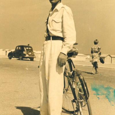 Il Vigile Umberto Tamburini in uniforme bianca e spallaccio nero con la bicicletta di servizio sul lungomare di Rimini. Sullo sfondo la spiaggia con le cabine in legno, una signora di spalle che attraversa la spiaggia e una topolino in transito 
