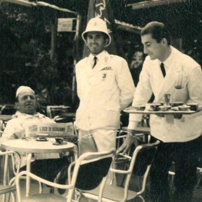 Il Vigile Umberto Tamburini in divisa bianca con casco coloniale, in piedi a lato di un tavolino di un bar, tra un cliente e il cameriere intento a servire 