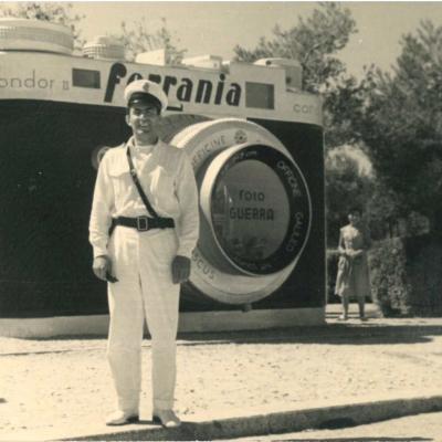 Il Vigile Corbelli in divisa bianca in posa davanti alla macchina fotografica nell'attuale piazzale Fellini. La macchina fotografica riporta il nome "Foto Guerra". Sullo sfondo, dietro le piante, la palazzina Roma. 
