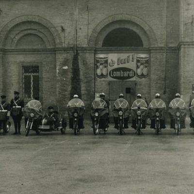 Foto di gruppo dei motociclisti dei Vigili Urbani nel 1959 in occasione di San Sebastiano. I Vigili sono schierati a lato del teatro Galli, sullo sfondo la pubblicità del supersucco Lombardi 