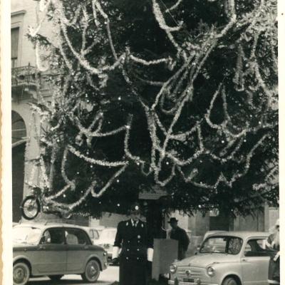 Immagine di piazza Cavour con l'albero di Natale posizionato davanti al Palazzo dell'Arengo. Davanti un Vigile Urbano con cappotto nero e cappello coloniale in occasione della Befana del Vigile.  