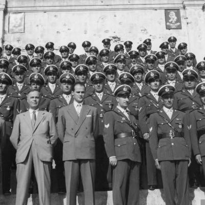 foto di gruppo dei Vigili Urbani di Rimini nel 1950. I Vigili sono disposti su delle scalinate su sie file, in fondo una parete con la pubblicità delle Vecchina Caffè. In prima fila, al centro, il Comandante Sanzio Della Marchina. 