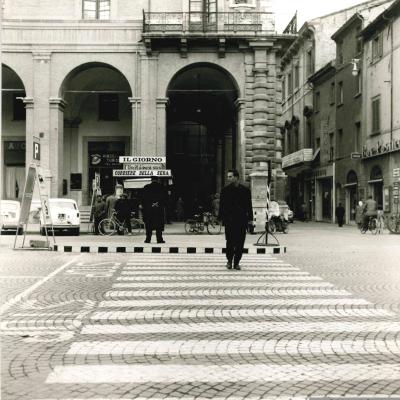 Attraversamento pedonale posto all'angolo di piazza Cavour corso d'Augusto davanti al Palazzo Garampi. è visibile un pedone intento ad attraversare la piazza e un Vigile Urbano di spalle che controlla un lato della strada. 