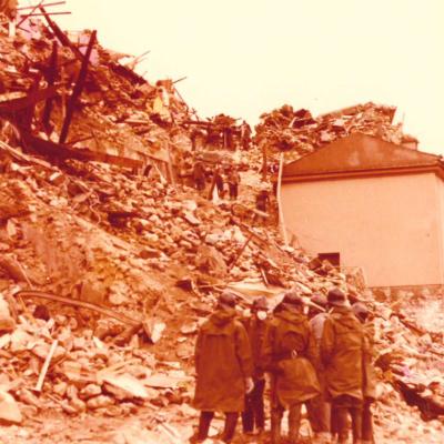 immagine di un gruppo di volontari in soccorso alle popolazioni civili in occasione del Terremoto del 1980, in piedi sopra un cumulo di macerie, sullo sfondo il muro di una casa ancora in piedi, tutto attorno case distrutte 