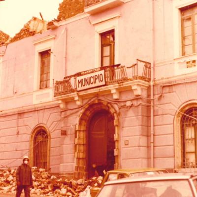 immagine del Municipio di Laviano distrutto dal terremoto del 1980. Ai lati della strada che passa davanti all'edificio, vi sono macerie. Sulla strada vi sono due auto ancora in sosta e un volontario che cammina 