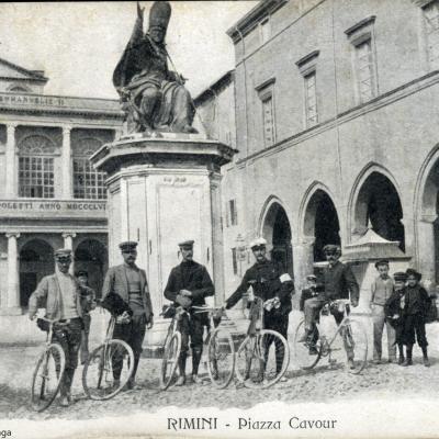riproduzione di una cartolina del 1911 in piazza Cavour con 6 Guardie Comunali con le loro biciclette, davanti alla statua di Paolo V, sul retro il teatro Galli a destra il palazzo del Podestà. Attorno a oro dei bambini