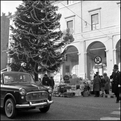 immagine di parte di piazza Cavour con un auto in primo paino e un Vigile che svolge servizio all'attraversamento pedonale. Sullo sfondo un albero di Natale con sotto i doni depositati in occasione della Befane del Vigile. Dietro il palazzo Garampi