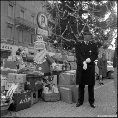 immagine di regali depositati in piazza Cavour sotto l'albero di Natale in occasione della Befana del Vigile. A lato dei regali il Vigile Aldo Trovanelli in alta uniforme. Sullo sfonfo l'insegna del Resto del Carlino