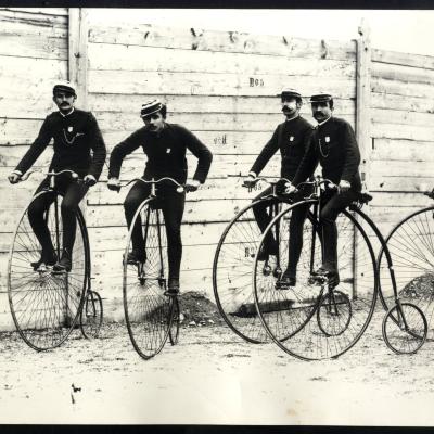 foto di cinque guardie comunali in divisa in sella alle biciclette di servizio. Le biciclette hanno la ruota anteriore molto grande mentre quella posteriore è piccola. Sullo sfonfo una staccionata in legno 